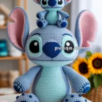 Two crocheted plush toys of a blue alien character with large ears; a smaller one sits on top of a larger one, surrounded by colorful yarn and crafting tools on a table, with sunflowers in the background.