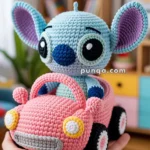 A crocheted toy featuring a light blue character with large ears and a pink nose, sitting inside a pink crocheted car held by a person's hand. The setting is indoors with a blurred background of shelves and a plant.