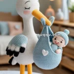A crocheted stork with a yellow beak holding a blue crocheted bundle, which contains a smiling baby doll wearing a matching blue hat, standing on a white table in a softly decorated room.