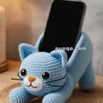 A blue crocheted cat-shaped phone holder with large eyes and an orange nose, holding a smartphone upright on a wooden surface.