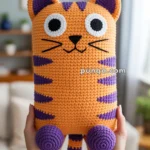 A person holding a crocheted cat pillow with an orange body and purple stripes and details, featuring large white and black eyes and a small nose, in a cozy living room setting.