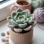 A crocheted succulent plant in a knitted pot is placed on a windowsill beside a cup of tea and several balls of yarn in pastel colors.