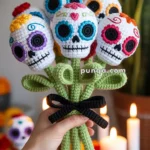 A hand holds a crocheted bouquet of sugar skull flowers, each with colorful decorations, surrounded by green crocheted leaves and stems tied with a black ribbon. In the background, there are blurred flowers and lit candles.