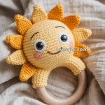 A crocheted toy in the shape of a smiling sun with a wooden ring attached, resting on soft fabric.