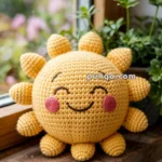 A crocheted sun toy with a smiling face, pink cheeks, and small sunray arms, sitting on a wooden windowsill beside green plants.
