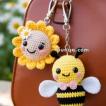 Two crocheted keychains, one depicting a smiling sun with a small flower and the other a cheerful bee with wings and a pink bow tie, attached to a brown handbag with a floral background.