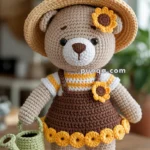 Crocheted teddy bear wearing a sun hat, brown overalls, and yellow sandals, holding a green watering can.