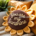 A crochet sunflower pillow with the words "You are my sunshine" is placed on a beige knitted blanket, surrounded by yellow cushions and greenery by a window.