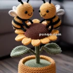 Two crocheted bees with smiling faces are sitting on a large crocheted sunflower in a crocheted pot, placed on a wooden floor with a sofa in the background.