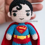 Crocheted keychain resembling a superhero, with a blue suit, red cape, and emblem on the chest, held in a person's hand.