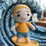 A crocheted doll wearing a yellow top and blue shorts stands on a crocheted yellow surfboard, with a knitted wave pattern in the background.
