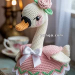 A crocheted swan figurine wearing a pink dress with green leaf details and a white bow, featuring a pink rose on its head. It is set on a wooden surface with a teacup and candle in the background.