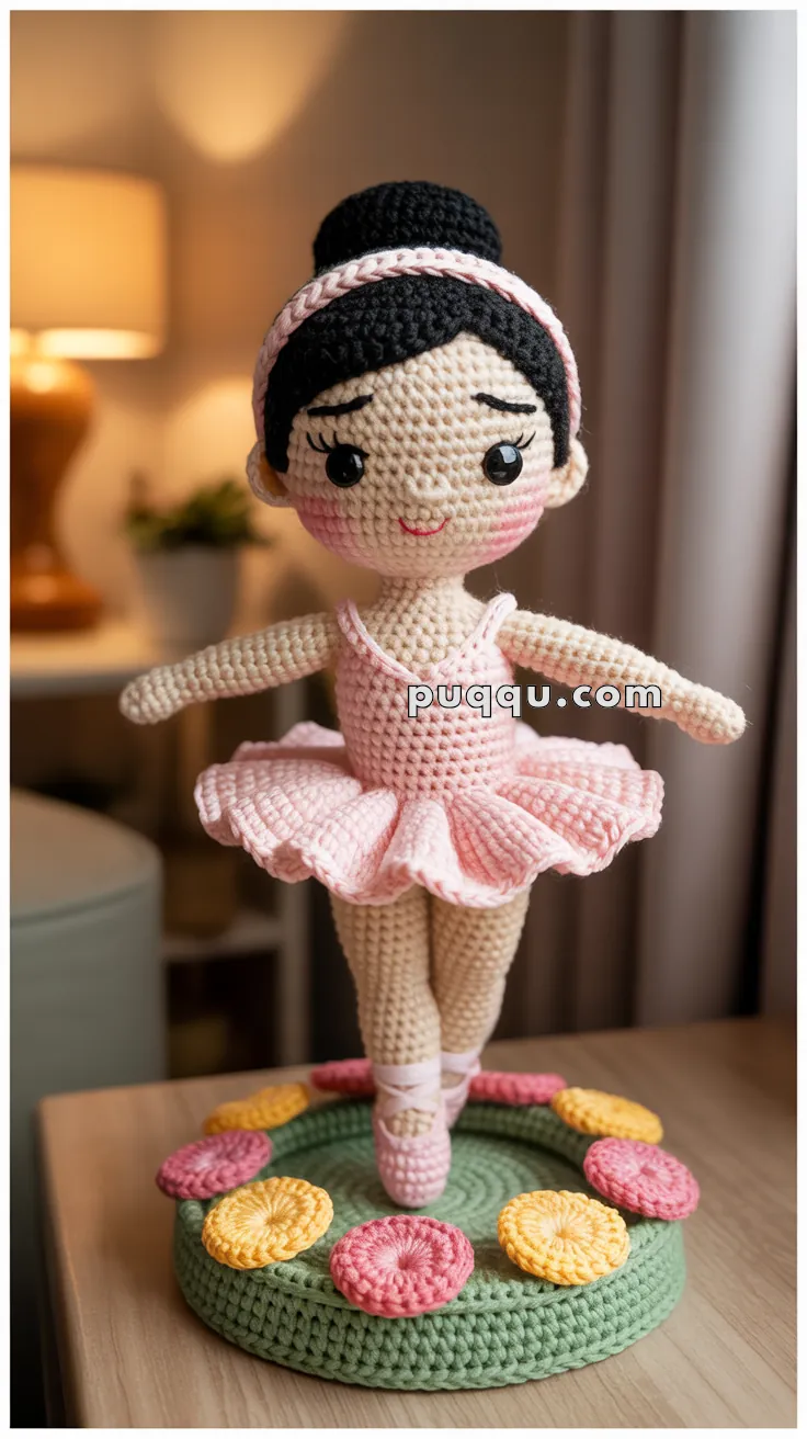 A crocheted doll in a ballet pose, wearing a pink tutu and matching ballet shoes, is displayed on a round crocheted base adorned with pink and yellow flowers.