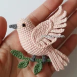 A small handcrafted macrame bird with pink feathers and black eyes, resting on a green branch, held in an open palm against a plain white background.