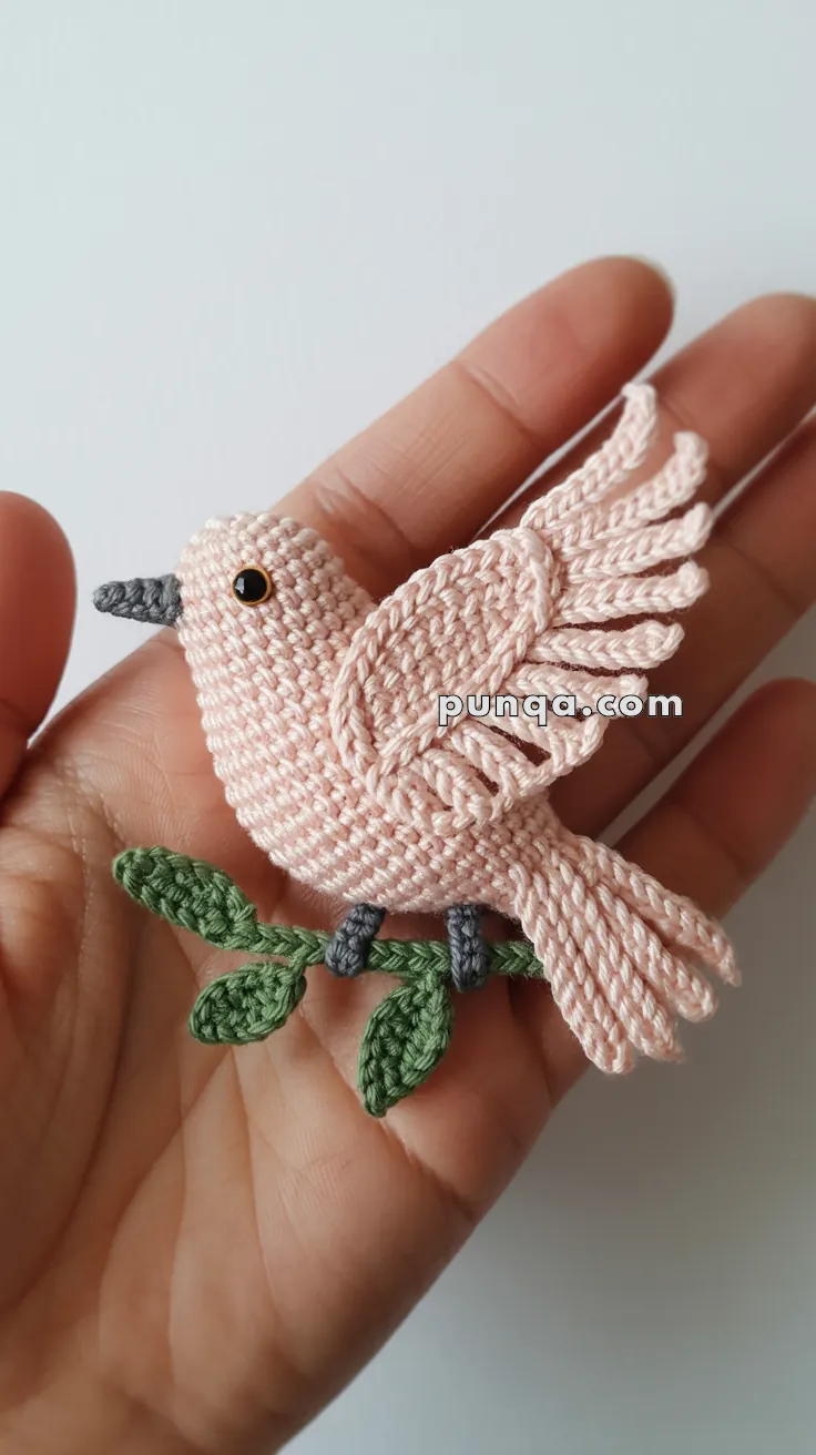 A small handcrafted macrame bird with pink feathers and black eyes, resting on a green branch, held in an open palm against a plain white background.