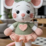 A crocheted amigurumi mouse with large ears and whiskers, wearing a green bow and a matching green shirt with a heart motif, along with a pink tutu skirt, standing on a wooden surface indoors.