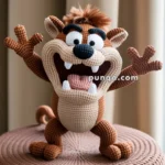 Crocheted plush toy resembling a cartoon Tasmanian Devil character with an open mouth and outstretched arms, sitting on a textured brown surface.