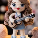 Crocheted doll with long brown hair, wearing a sparkly top and a blue skirt, holding a guitar. The doll is standing on a wooden surface with yarn balls and a crochet hook nearby, and a crocheted microphone on a stand in front.