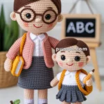 Two crocheted dolls, an adult and a child, stand on a circular mat. The adult doll wears glasses, a pink sweater, and a gray skirt, holding a yellow book. The child doll wears a gray skirt, white shirt, and holds a pencil. They are positioned in front of a miniature blackboard with "ABC" written on it. Small crochet items, including an apple and letter blocks, are scattered on the mat.