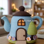 A cute crocheted teapot cozy designed to look like a small house, featuring a blue base with a round brown roof, an arched window, a door, and themed decorations including a tree and flowers. It is placed on a wooden table in a cozy, softly-lit room.