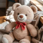 A crocheted teddy bear with a beige body, white paws and muzzle, and a red bow tie, sitting on a fabric surface near a window with books in the background.