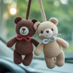 Two crocheted teddy bears, one brown and one beige, hanging from the rearview mirror of a car, each adorned with a small bow tie, against a blurred green background.