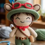 Crocheted doll with a green hat, red glasses, blue feather, and brown bag, smiling with closed eyes on a wooden table.