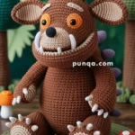 A handmade crocheted brown creature with big round ears, orange eyes, and a wide mouth displaying white tusks sits on a green mat, surrounded by a forest-like setting with crocheted trees and mushrooms.