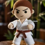 A crocheted martial arts doll with a brown belt and brown hair stands in a fighting stance on a wooden surface, with plants in the background.