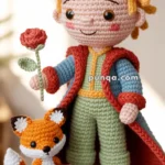 A crochet doll resembling a young character with yellow hair, holding a red flower, and wearing a red and blue attire. A small crocheted fox stands beside the doll on a blue base with star decorations.
