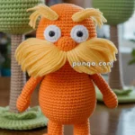 A handmade crocheted orange figure with large yellow eyebrows and mustache, featuring prominent eyes, standing on a wooden table. Behind it are green crocheted trees and blurred string lights in the background.