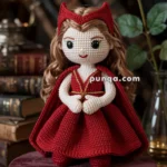A handmade crochet doll with long brown hair, wearing a red dress and matching cape, standing on a round, velvet cushion; the background features a rose and stacked books.