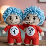Two crocheted dolls with blue curly hair wearing red outfits labeled 'Thing 1' and 'Thing 2,' positioned on a wooden surface in a room with a soft pink background.