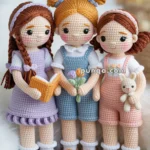 Three crocheted dolls standing together, each with distinct hairstyles, holding a book, flowers, and a bunny toy, dressed in pastel clothes and placed on a green knitted surface.