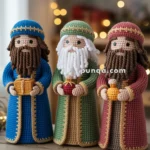 Three crocheted figurines representing the Three Wise Men, each holding a gift and wearing intricately designed robes in blue, green, and red colors, are displayed on a wooden surface with blurred festive lights in the background.