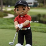 A crochet doll of a golfer wearing a red shirt, black pants, a black cap, and holding a golf club, standing on a golf course.
