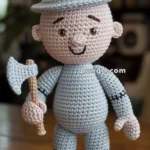 A crocheted doll resembling a tin man, wearing a pointed hat and holding a small axe, is placed on a wooden table with a blurred background of shelves and a plant.