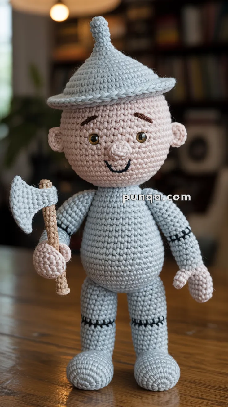 A crocheted doll resembling a tin man, wearing a pointed hat and holding a small axe, is placed on a wooden table with a blurred background of shelves and a plant.
