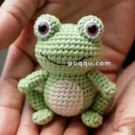 A small, crocheted green frog with large eyes and a white belly held in someone's hands.