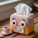 A crocheted tissue box cover with cartoon-like eyes and a sign saying "Cry it out, I'm here" on a wooden table. Beside it is a small crocheted pink teacup.