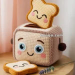 Crocheted toaster and bread plush toys with cute facial expressions, placed on a wooden table next to a purple flower and pink curtains in the background.