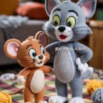 Crocheted plush toys of a gray cat and a brown mouse stand on a checkered rug, surrounded by yarn balls and a small toy cheese.