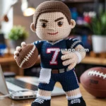 A crocheted doll dressed as a football player in a blue jersey with the number 7, holding a football, standing on a table in a room with a laptop and books in the background.