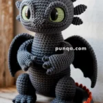 A crocheted dragon toy with large green eyes and black wings, sitting on a wooden surface with a red-tipped tail.