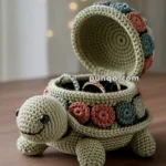 A crochet turtle-shaped box with an open shell displaying rings inside, featuring a smiling face and colorful patches.
