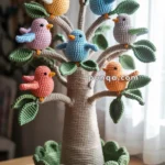 A crocheted tree with multicolored amigurumi birds perched on its branches, featuring green leaves and a round base, placed on a wooden surface near a window with soft light.