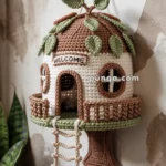 Crocheted treehouse wall hanging with a round brown roof adorned with green leaves, a "Welcome" sign above the door, and a rope ladder leading to a porch.