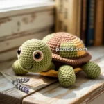 A crocheted turtle toy with a green head and legs, a brown and yellow shell, sitting on a wooden table beside two sprigs of lavender, with books blurred in the background.