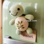 A crocheted turtle magnet with a smiley face and embroidered shell design on a pastel green refrigerator door, accompanied by a small card labeled "Shelbert."