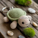 A crocheted turtle keychain with a green shell and beige body, placed on a wooden surface surrounded by small stones and moss balls.